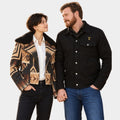 A man and a woman standing side by side against a light gray studio background. The woman wears a black-and-tan geometric patterned jacket with a black shearling-style collar over a white shirt and black jeans.