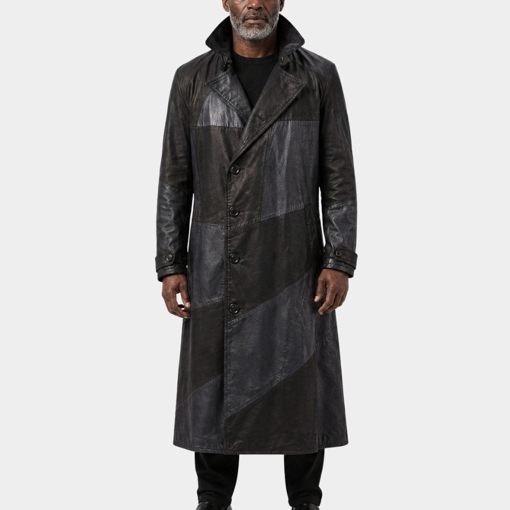 A man wearing a long black leather coat with diagonal paneling, photographed against a plain light gray studio background.